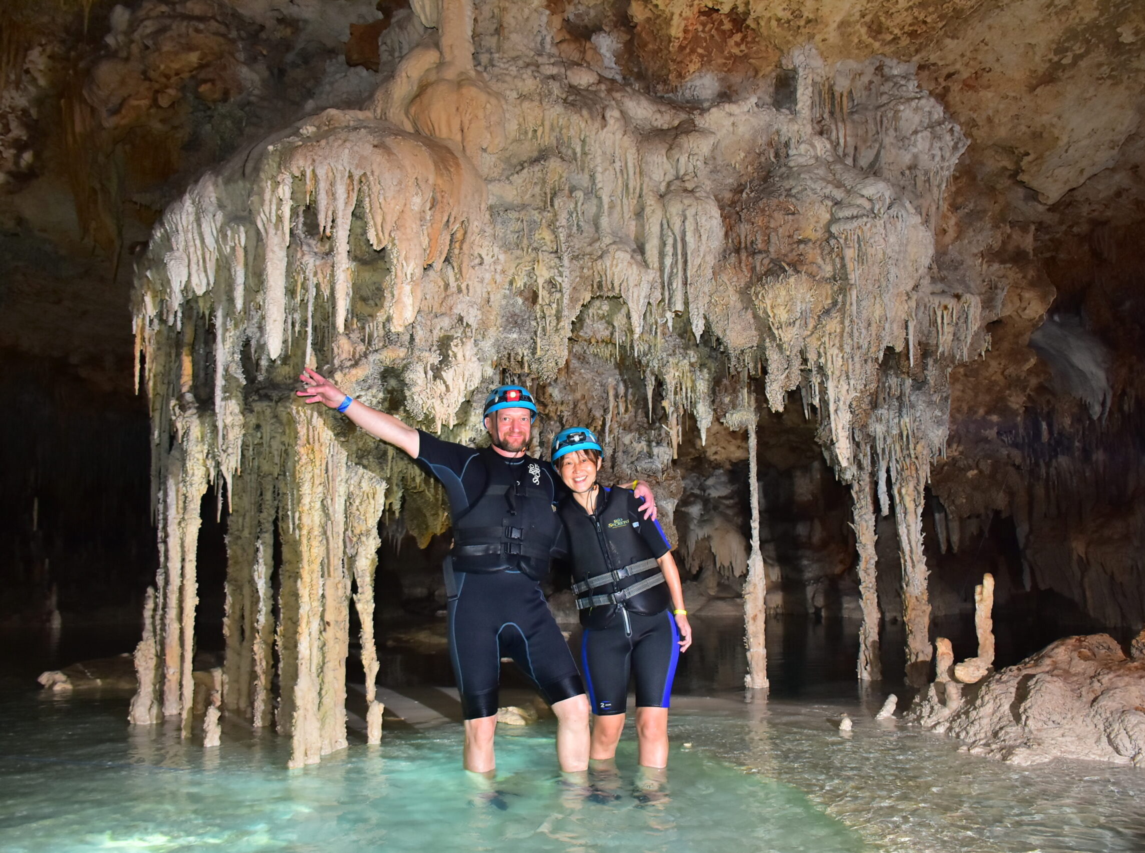 Stefan Motz and Yoko San exploring Rio Secreto’s underground river cave, wearing helmets and waving amidst limestone formations.