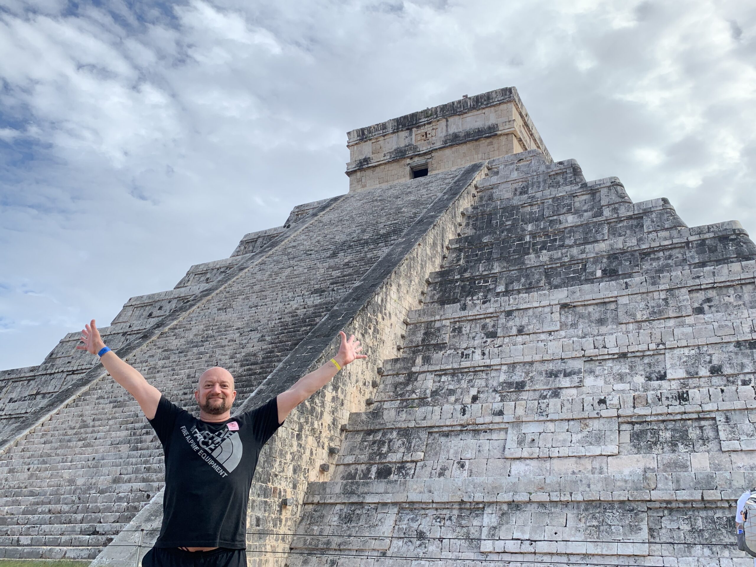 Stefan Motz smiling with outstretched arms under the sunny sky at Chichen Itza, Yucatan Peninsula, Mexico, with clouds above the ancient Maya pyramid.